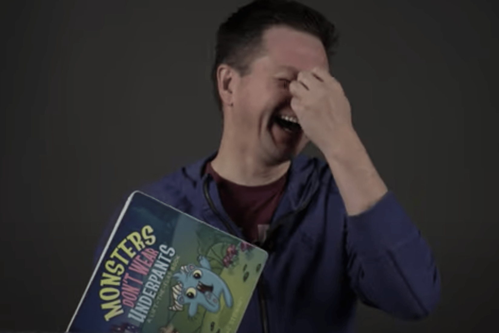 Author Isaac Stewart covering his face and laughing while holding a copy of "Monsters Don't Wear Underpants."